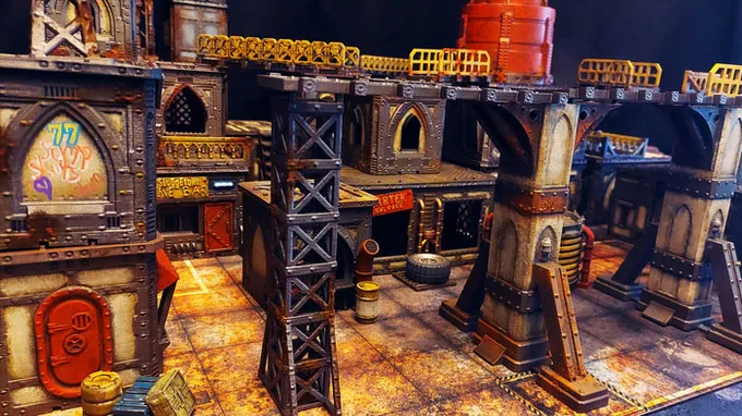 A detailed tabletop scene showcases a grimdark, sci-fi urban environment with various structures made from textured materials, featuring rusted metal and weathered surfaces. Prominent elements include tall support beams, stacked containers, and buildings adorned with graffiti and signage, all set on a gritty, tiled ground. Elevated walkways connect the structures, enhancing the industrial feel of the UnderNidus District.