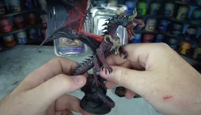 A detailed dragon miniature is being held in a hand, showcasing its intricate features, including prominent scales, fierce facial expression, and large wings with a red underside. The dragon is painted in dark hues with highlights that accentuate its muscular form and menacing appearance. In the background, various paint jars are visible, indicating a workspace for model painting.