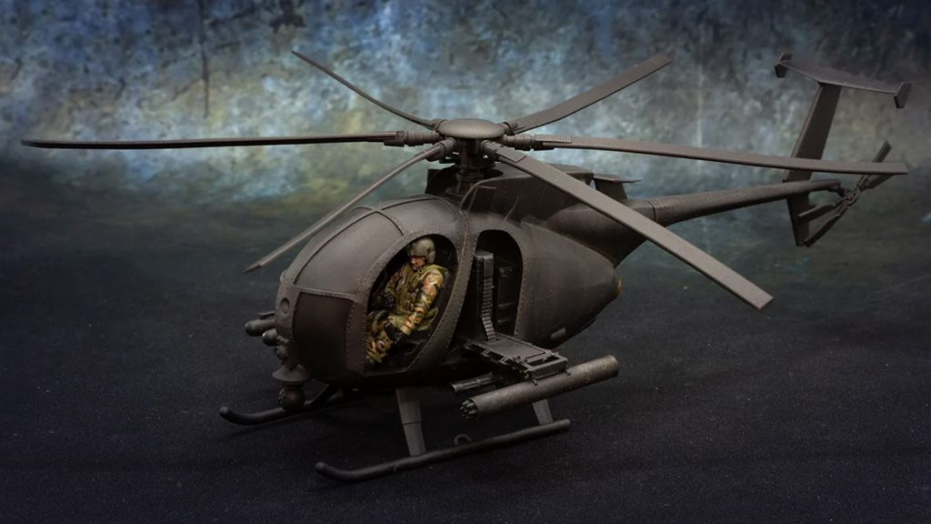 A detailed model of a Little Bird helicopter, specifically the AH-6 variant, is prominently displayed. The helicopter features a sleek, dark exterior and is equipped with a mounted weapon system on the side. Inside the cockpit, a miniature figure dressed in camouflage is seated, showcasing the model's intricate design and attention to detail. The background is a textured gradient, enhancing the focus on the helicopter.