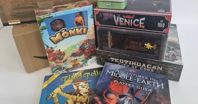 Stack of board games and books, including "MØNKI", "VENICE", "Adventures in Middle-Earth", and "Fiend Folio".