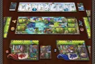 A vibrant tabletop scene showcases the board game Mystic Lands, featuring a central game board adorned with colorful illustrations of landscapes, creatures, and various resource tracks. Surrounding the main board are player boards, each displaying unique artwork and spaces for cards and tokens, along with dice and resource cubes in different colors. The layout includes a deck of cards at the top, with additional components like tokens and a score tracker, all set against a rich, textured background.