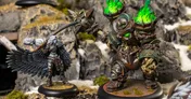 A detailed miniature scene features a character with large, intricately designed black wings, dressed in a mix of armor and clothing, wielding a weapon and casting a spell. Opposite this character stands a massive, armored figure with glowing green accents and a spiked weapon, set against a rocky terrain with patches of moss. The contrasting sizes and designs of the two figures highlight the dynamic nature of the scene.
