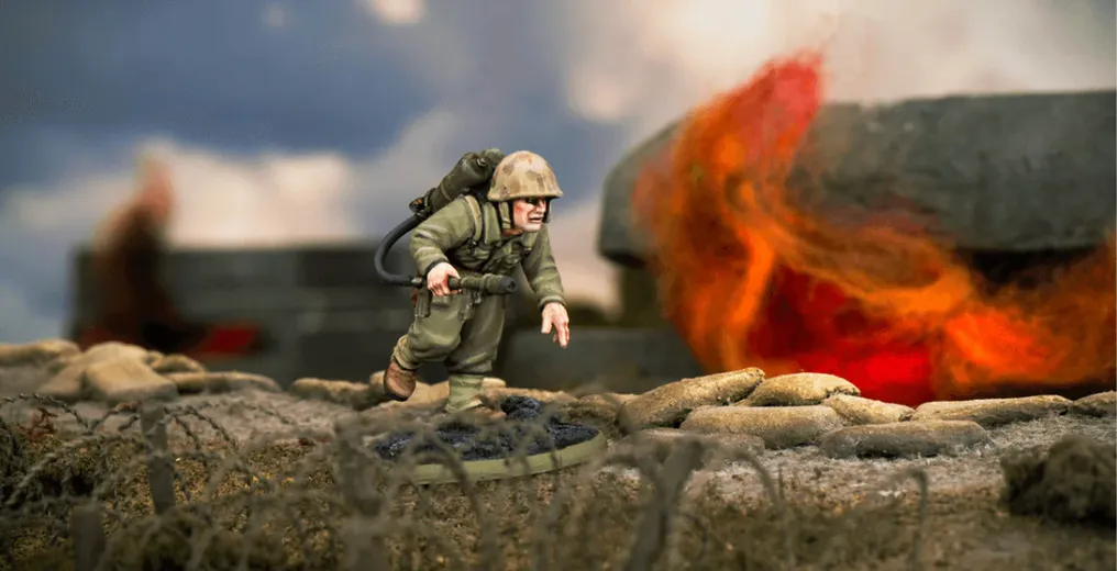 A detailed miniature of a U.S. soldier, Hershel W. 'Woody' Williams, is depicted in action, equipped with a flamethrower on his back and wearing a helmet and military uniform. He is positioned on a textured base that resembles a battlefield, with a backdrop of flames and smoke suggesting a combat environment. The scene captures the intensity of the Pacific theatre during World War II.