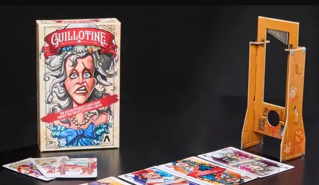 The image features the box of the card game "Guillotine," prominently displaying an illustration of a startled woman with gray hair and a blue bow, set against a light-colored background. Next to the box, a wooden guillotine prop is shown, designed to hold the noble cards. In the foreground, several tarot-sized noble cards are laid out, showcasing colorful illustrations of various characters, each with distinct expressions and attire, reflecting the game's theme of the French Revolution.