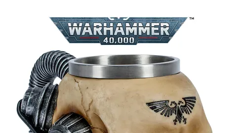 Beige ceramic mug shaped like a stylized human skull with mechanical augmentations. A metal rim lines the top of the mug. Two large, cylindrical, metallic mechanical components resembling pistons or servos are attached to the sides of the skull, connected by coiled black cables. A black eagle emblem with outstretched wings and a skull in the center is tattooed or printed on the forehead. The background shows a metallic banner with the text "WARHAMMER 40,000" in bold, futuristic lettering. The banner has a red and white stripe along the bottom edge.