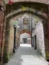 A stone archway leads into a narrow passageway within a medieval castle, featuring weathered red stone walls and a cobblestone floor. Colorful triangular flags hang above, adding a festive touch, while soft lighting from wall-mounted fixtures illuminates the scene. In the distance, an open doorway reveals a glimpse of the castle's interior, enhancing the atmospheric and slightly haunted vibe of the setting.