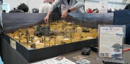 Hands adjusting miniatures on a wargaming diorama with a stone house, trees, and a tank.
