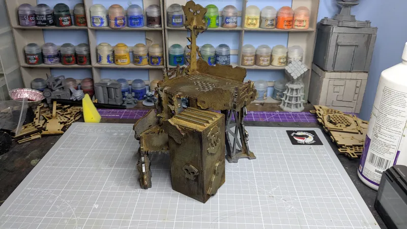 An Orc Lookout structure is prominently displayed, featuring a multi-level design with a weathered, rustic appearance. The lookout has a large, central platform with a grid-like surface, supported by a framework of beams and a sturdy base that includes a door adorned with a skull motif. In the background, shelves filled with various paint pots and miniature components are visible, indicating a workspace dedicated to tabletop gaming.