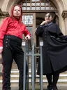 Two individuals pose in front of a grand, gothic-style building. One person, wearing a bright red shirt and black trousers with red stripes, stands confidently with arms crossed. The other person, dressed in a long black coat with a high collar and a blue shirt underneath, holds the hem of their coat, creating a dramatic silhouette. The ornate entrance of the building, featuring large wooden doors and intricate stonework, serves as a striking backdrop.
