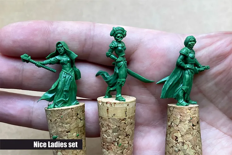 Three green resin miniatures are displayed on cork bases, held in a hand. The figures represent fantasy female characters: on the left, a warrior wielding a mace and wearing a flowing robe; in the center, a character with a sword and a distinctive headdress; and on the right, another figure in a cloak. The text 
