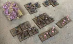 A collection of Zayken miniatures from Void Scar Miniatures is displayed on a textured surface. In the foreground, several bases feature hordes of zombified figures in various colors, including green, purple, and pink, representing the Neophyte Mobs. To the left, a tank is adorned with grotesque, bloated creatures, suggesting a fusion of machine and undead, while additional bases of zombies are scattered throughout the scene, emphasizing the overwhelming numbers of the Zayken forces.