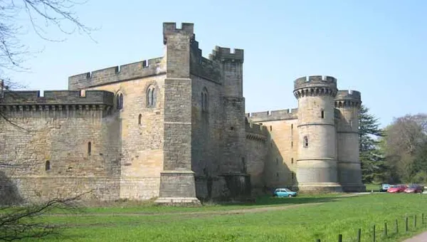 A large stone castle with multiple towers and thick walls, set against a clear blue sky. The castle features a mix of architectural styles, with rounded towers and crenellated parapets. In the foreground, a grassy area is bordered by a low fence, and a few cars are parked nearby.