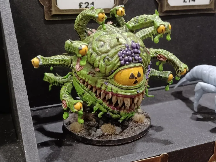 A vibrant green, multi-eyed monster with a large, toothy grin and several tentacle-like appendages is displayed on a circular base. The creature features a prominent yellow eye with a radioactive symbol, surrounded by purple and green details, and is set against a dark background. In the foreground, small tufts of grass and rocks add to the diorama's texture, while a smaller, light blue figure is partially visible to the right.