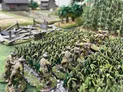 A group of miniature soldiers in khaki uniforms is advancing through dense green foliage, with some figures holding rifles and others appearing to be in various poses of movement. The background features a detailed landscape with palm trees and a rustic building, enhancing the immersive setting of the wargaming scene. The terrain is richly textured, with a mix of grass and underbrush, creating a realistic battlefield environment.
