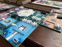 Board game tiles showing a scenic route with blue water, green hills, and white snowflake-like structures. Miniature buildings and a bridge are visible. Text on tiles includes "Little Rocket Games" and "Nakasendo."