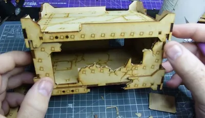 A partially assembled, light brown cardboard or MDF terrain piece with a damaged, jagged top edge, revealing an open interior. The structure has square and rectangular cutouts, with small square and circular holes along the edges. It rests on a green self-healing cutting mat with a grid pattern. A hand is visible on the left side, holding part of the terrain. Small debris and tools, including a metal L-shaped piece, are scattered on the mat.