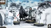 A detailed scene from Star Wars Legion features iconic miniatures set in a snowy landscape. Prominently displayed are Darth Vader, holding a red lightsaber, and a uniformed officer pointing, surrounded by several Stormtroopers. The terrain includes patches of grass and rocky elements, enhancing the immersive atmosphere of the battlefield.