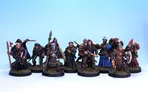 A diverse group of painted miniatures stands against a light blue background, showcasing various adventurers and characters. The figures include a mix of warriors, spellcasters, and rogues, each detailed with unique clothing, weapons, and expressions. They are mounted on circular bases with tufts of grass, highlighting their fantasy theme and adding to the overall aesthetic of the collection.