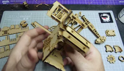 A partially assembled oil pump model made from laser-cut MDF is being held in hands, showcasing intricate details like vents and supports. Surrounding the model are various unassembled MDF pieces, indicating ongoing construction. A cutting mat with a grid pattern serves as the work surface, and a sticker reading "Orc Miniatures Hate Frogs" is visible in the background.