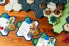 A tabletop gaming scene features a modular hex terrain setup with various textured tiles in shades of green and gray. In the foreground, several character cards are displayed, each with colorful artwork and stats, including figures like Duke and Scarlett. Miniatures representing characters from the G.I. JOE universe are positioned on the terrain, ready for gameplay, alongside red wound markers and other game components.