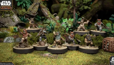 A group of four Ewok miniatures from the Star Wars: Legion game is displayed on circular bases, set against a lush, green forest backdrop. Each Ewok is uniquely posed: one holds a spear, another brandishes a slingshot, while the others are equipped with various weapons and accessories. The terrain features rocks, foliage, and remnants of a crashed vehicle, enhancing the scene's immersive quality.