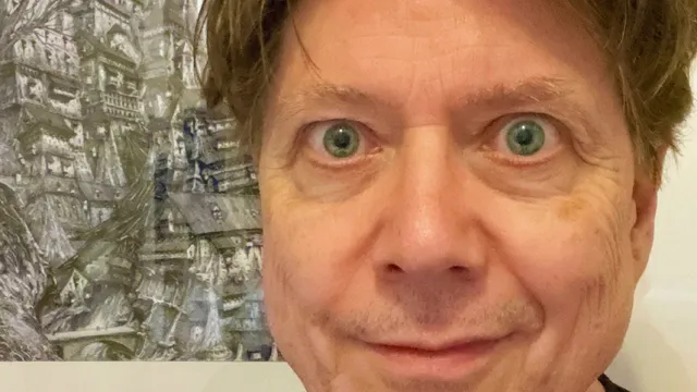 A close-up portrait of a man with light brown hair and striking green eyes, smiling slightly. He is wearing a black shirt and is positioned in front of a detailed, artistic background featuring a complex, fantastical structure, possibly a building or landscape. The overall tone of the image is casual and inviting, with a focus on the subject's expressive features.