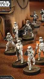 A group of Star Wars miniatures features several Stormtroopers in white armor, positioned in a desert-like environment with a backdrop of a weathered building. Among them stands a prominent figure in black armor, holding a red lightsaber, likely representing Darth Vader. The scene is detailed with various poses of the troopers, some aiming weapons, while others appear to be in action, all set on circular bases with textured surfaces.