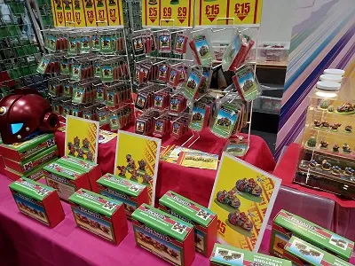 A vibrant display of miniature wargaming products is showcased on a table covered with a bright pink cloth. Various boxes of miniatures are arranged neatly, with some priced at £9 and others at £15, featuring colorful artwork on their packaging. Behind the boxes, additional miniatures are displayed in clear packaging, highlighting a diverse selection for wargaming enthusiasts.