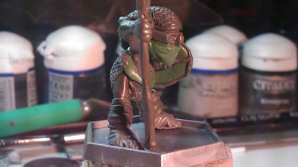 A small, detailed miniature figure stands on a square base, holding a staff and wearing a green mask. The figure is dressed in textured armor, showcasing intricate details like scales and straps. In the background, various paint pots and tools are visible, indicating a workspace for painting or modeling miniatures.