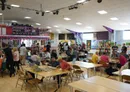 A spacious gaming hall filled with tables and chairs, where groups of people are engaged in various tabletop games. In the background, shelves are stocked with board games, and a vibrant display area features colorful banners and decorations. Attendees are seen socializing and playing games, creating a lively and inclusive atmosphere.