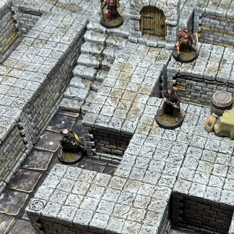 A detailed tabletop scene features modular dungeon blocks made of textured stone, arranged to create walls, platforms, and staircases. Several miniatures, including two armored figures and a robed character, are positioned on the blocks, exploring the dungeon layout. A wooden barrel and a small pile of supplies are visible, adding to the immersive environment.