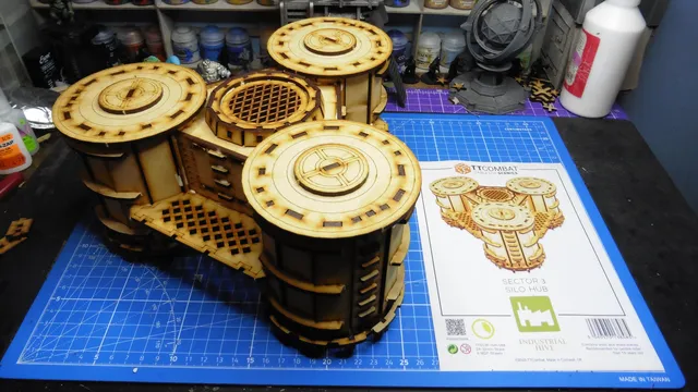 A detailed model of the Sector 3 Silo Hub from TT Combat is prominently displayed on a blue cutting mat. The structure features multiple cylindrical silos with intricate wooden textures and circular tops, showcasing a combination of open latticework and solid panels. In the background, various paint pots and a few other miniature models are visible, along with the instruction sheet for the Silo Hub, which includes an illustration of the assembled model.