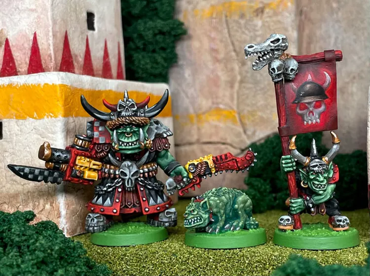 A trio of space orcs stands prominently in the foreground, featuring Attila the Warlord, who is adorned in elaborate armor with spikes and a large horned helmet, wielding a futuristic weapon. Beside him is a small green orc hound, Snaarl, and Groblett the Goblin, who holds a banner displaying a skull and horns. The background includes stylized stone structures and greenery, enhancing the fantastical setting.