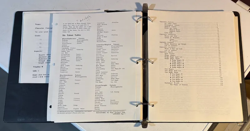 A close-up view of a three-ring binder containing pages from the playtest manuscript for "ManaStorm: The Magic Adventure Game." The visible pages include character concept sheets with sections for character name, goals, aspects, vitality, and life. Additionally, there are tables listing various talents associated with different colors of magic, such as Black, Blue, Red, and Green, along with their prerequisites. Handwritten notes are visible in the margins, indicating additional thoughts or clarifications.