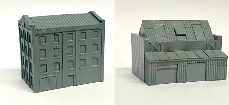 A detailed model of an industrial building designed for 6mm scale wargames, featuring a textured brick facade and multiple windows. The structure has a flat roof with visible paneling and a large door on the front, along with smaller doors and windows on the sides. The model is cast in a gray resin, showcasing its intricate design suitable for historical or modern warfare settings.