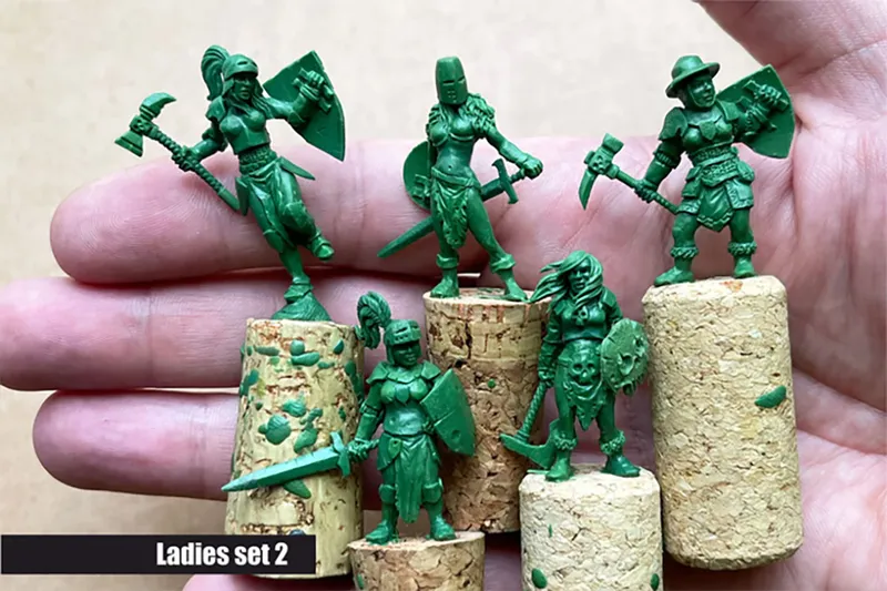 A hand holding several green resin miniatures from the "Ladies Only" collection by Scibor Monstrous Miniatures. The miniatures depict various fantasy female characters, including a warrior with an axe and shield, a knight, and a pirate, all standing on cork bases. The text "Ladies set 2" is displayed at the bottom of the image.