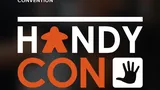 White text on dark background reads "UK BOARD GAME CONVENTION." Large orange and white text spells "HANDYCON" with an orange icon of a person in a wheelchair between letters. To the right, an orange hand icon. Small white text at bottom reads "DAVENTRY, UK."
