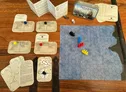A compact naval wargame setup features the "Battle Box - Age of Sail" tin, which contains various game components. The scene includes a hexagonal grid battle mat with a blue ocean design, four colored ship tokens representing different vessels, and tracking tokens. Nearby are cards for ship status, critical effects, and scenarios, along with dice and a rule booklet, all arranged on a wooden surface.