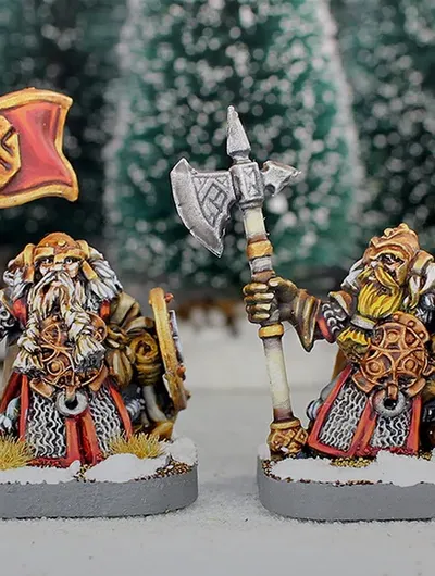Three painted miniature figures of fantasy dwarves stand on round bases in a snowy outdoor setting with blurred evergreen trees in the background.

The left figure wears a horned helmet, patterned tunic, and fur cloak, holding a pole-mounted red banner with a gold emblem of a snarling face. The figure also carries a round shield on their back.

The center figure, slightly behind the others, wears a helmet with a nose guard, a fur-lined cloak, and carries a pole weapon with a curved blade.

The right figure, holding a large double-bladed axe over one shoulder, wears a horned helmet, fur cloak, and patterned tunic. Their beard is braided and wrapped around their waist.