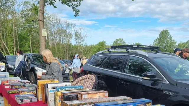 Outdoor car boot sale with red tablecloths displaying board games, DVDs, and books. People browsing and parked cars in background.