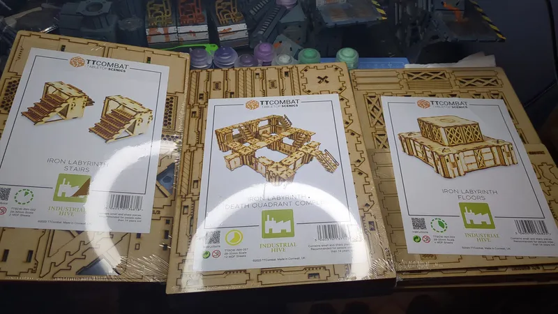 Two packages of TT Combat tabletop scenery are displayed on a work surface. The left package is labeled "Iron Labyrinth: Death Quadrant Complex," featuring a design of interconnected structures. The right package is labeled "Iron Labyrinth: Floors," showcasing a stack of storage tanks. In the background, various paint pots and additional terrain pieces are visible, indicating an active hobby workspace.