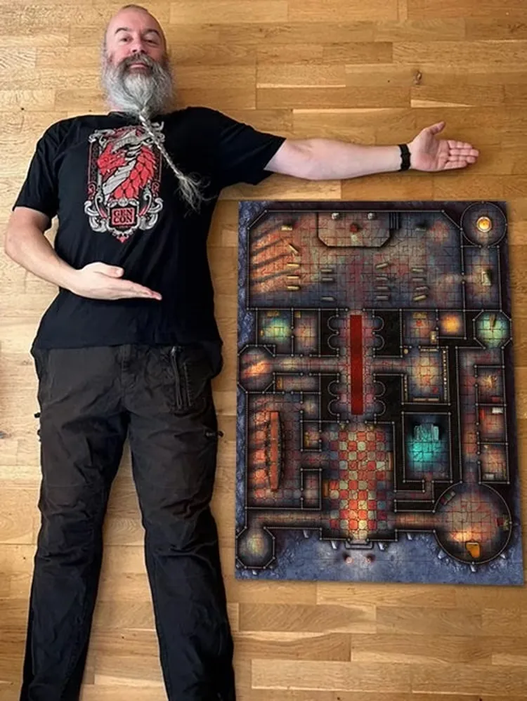 A person lies on a wooden floor next to a large, detailed dungeon map, which features various rooms, corridors, and colorful lighting effects. The map includes a grid layout with distinct areas marked by different textures and colors, such as red, blue, and green. The individual is wearing a black t-shirt with a graphic design and has a long beard, extending one arm towards the map to indicate its size.