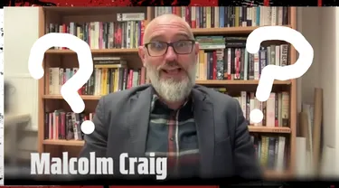 A man with a beard and glasses, dressed in a plaid shirt and blazer, is smiling and gesturing enthusiastically. He is seated in front of a bookshelf filled with various books, suggesting a scholarly environment. The image features large question marks overlaying his face, adding a playful or mysterious element to the scene. The background has a dark, textured border, enhancing the overall dramatic effect.