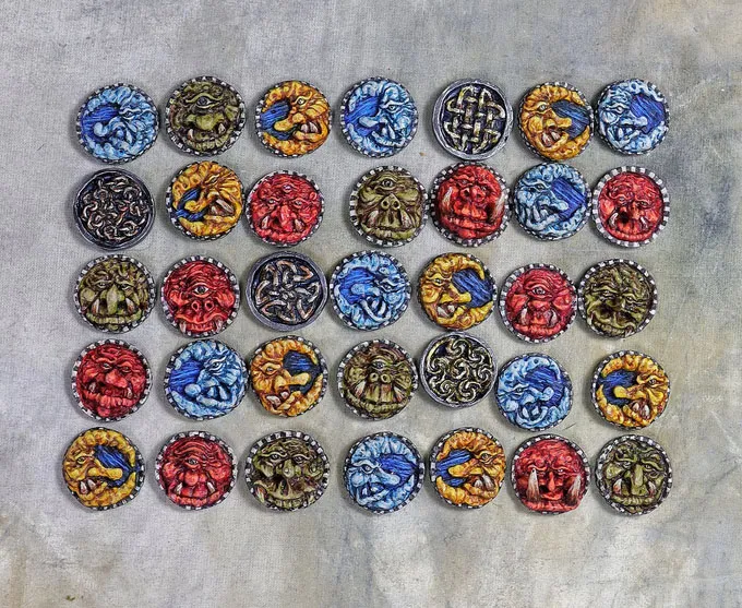A grid of 36 hand-sculpted shields, each featuring intricate designs inspired by Celtic mythology. The shields display a variety of colorful motifs, including scowling faces, gurning expressions, and detailed knotwork, rendered in vibrant hues of red, blue, green, yellow, and black. The background is a neutral gray, allowing the vivid colors and intricate details of the shields to stand out prominently.