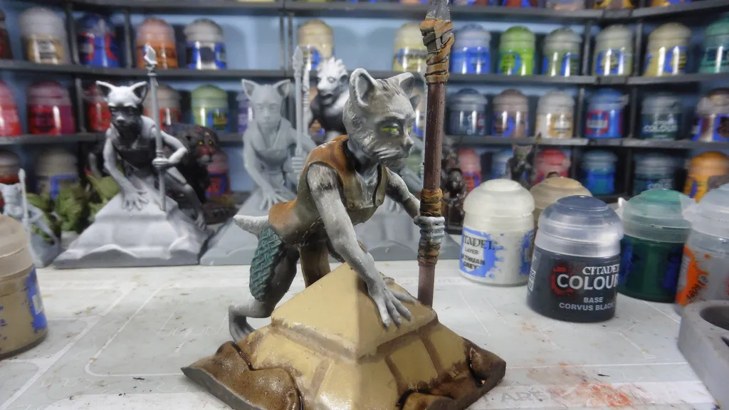Miniature figurine of a feline humanoid with green eyes, brown fur, and a brown vest, kneeling on a pyramid base. Staff with a crystal top in right hand. Nearby: two paint pots, one labeled "Citadel Colour Base Corvus Black." Background shelves hold more figurines and supplies.