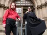 Two individuals pose in front of a grand, gothic-style building. One person, wearing a bright red shirt and black trousers with red stripes, stands confidently with arms crossed. The other person, dressed in a long black coat with a high collar and a blue shirt underneath, holds the hem of their coat, creating a dramatic silhouette. The ornate entrance of the building, featuring large wooden doors and intricate stonework, serves as a striking backdrop.