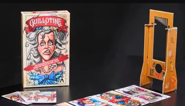 The image features the box of the card game "Guillotine," prominently displaying an illustration of a startled woman with gray hair and a blue bow, set against a light-colored background. Next to the box, a wooden guillotine prop is shown, designed to hold the noble cards. In the foreground, several tarot-sized noble cards are laid out, showcasing colorful illustrations of various characters, each with distinct expressions and attire, reflecting the game's theme of the French Revolution.