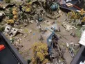 A detailed tabletop gaming diorama showcases a rugged, rocky terrain with scattered ruins and overgrown vegetation. The landscape features a mix of rocky outcrops, broken structures, and patches of yellow foliage, creating a post-apocalyptic atmosphere. Small miniatures are positioned throughout the scene, suggesting an ongoing game or battle among the ruins.