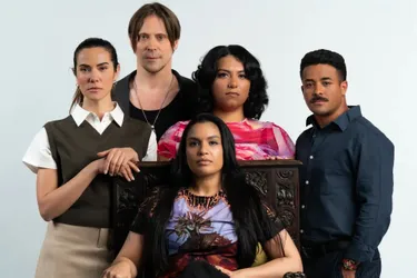 A group of five individuals poses together, with a mix of expressions and styles. The central figure, seated, wears a colorful shirt featuring a dragon design, while the others stand around, showcasing a variety of outfits, including a dark vest and a patterned blouse. The background is a simple light color, allowing the group's diverse looks and the ornate chair they are gathered around to stand out.