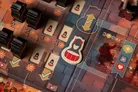 Board game with devil-themed tiles, numbered cards showing icons of drummers, devils, and fireworks. Central tile features a devil icon with red belt. Arrows indicate movement or action direction. Numbers "3" and "2" visible on cards.